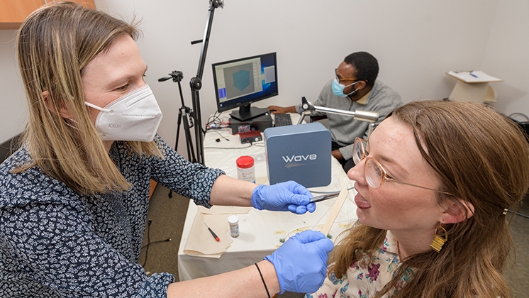 A researcher in a lab wearing a mask and blue latex gloves collects a sample from a research subject's mouth while another researcher records data on a desktop computer.