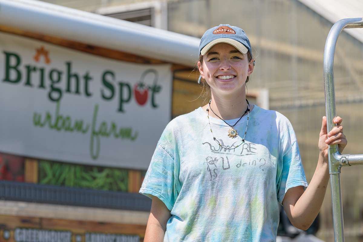 University of Delaware female student wearing baseball cap and blue tie-dye t-shirt stands in front of  sign reading 