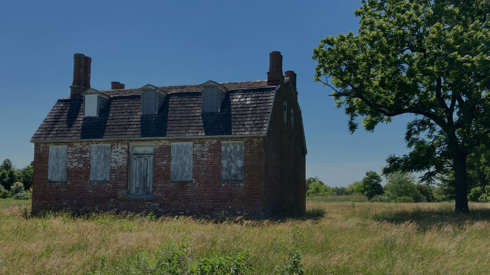 Photo of an abandoned brick structure with boarded up windows surrounded by a bright blue sky, grass and trees