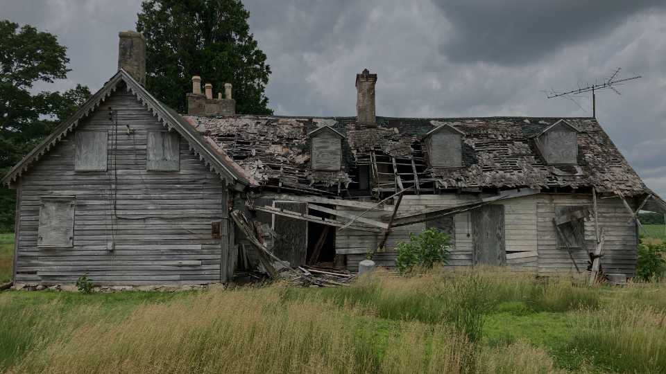 Photo of a dilapidated, older home with boarded up windows and partially collapsed wooden siding, surrounded by a cloudy grey sky and grass