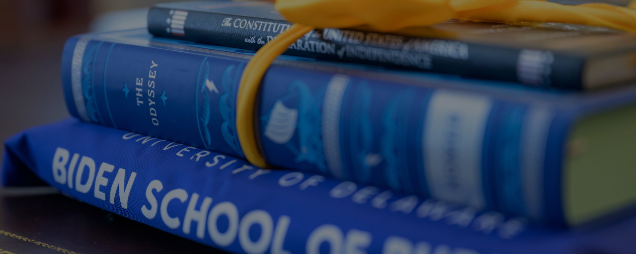 Photo of a blue Biden School t-shirt folded and stacked beneath a hardback copy of The Odyssey by Homer and a bound copy of the U.S. Constitution