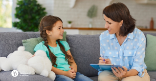 A mother holding a tablet smiling and talking to her daughter on the couch.