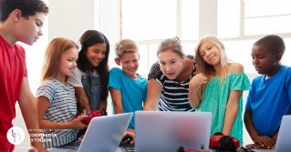 A group of kids around a table with a teacher looking at laptops