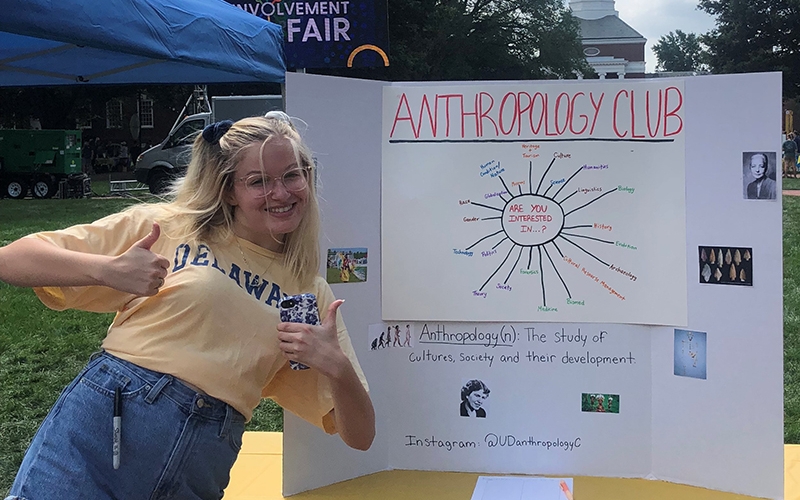 A University of Delaware student wearing a. yellow "Delaware" shirt makes the "thumbs up" sign while standing next to a sign for the Anthropology Club on the Green at UD during the fall Involvement Fair.