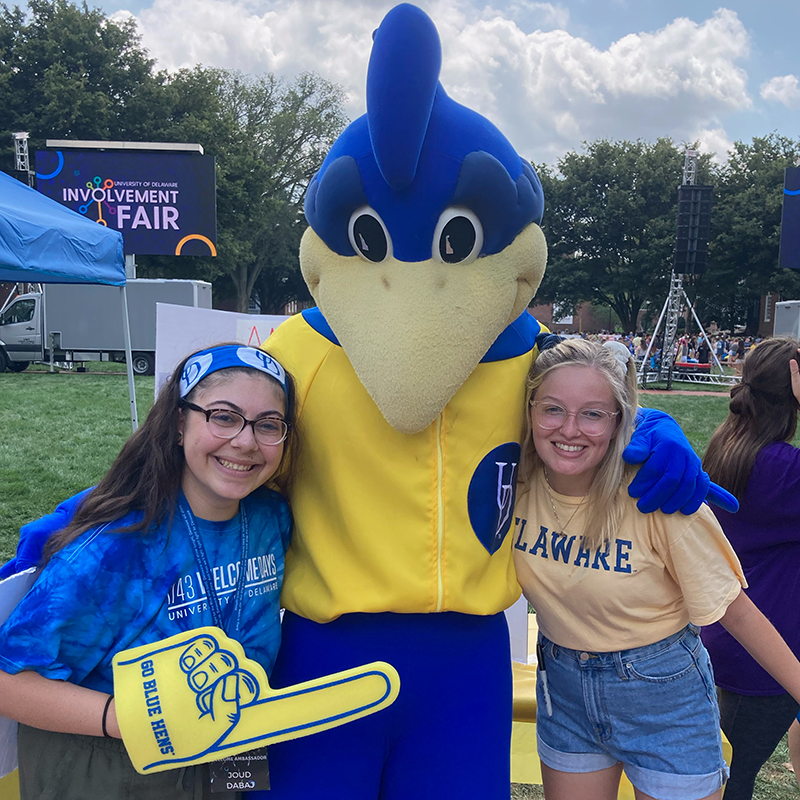 Two University of Delaware students dressed in spirit wear pose for a picture the YoUDee the official mascot, during the fall Involvement Fair on the Green.