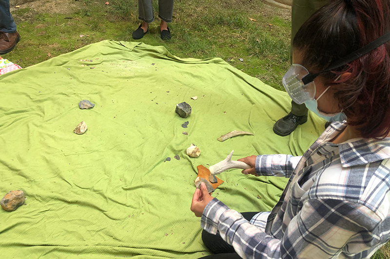 A University of Delaware student and Anthropology Club member practices making stone tools on a green blanket, using an antler to shape a flint.