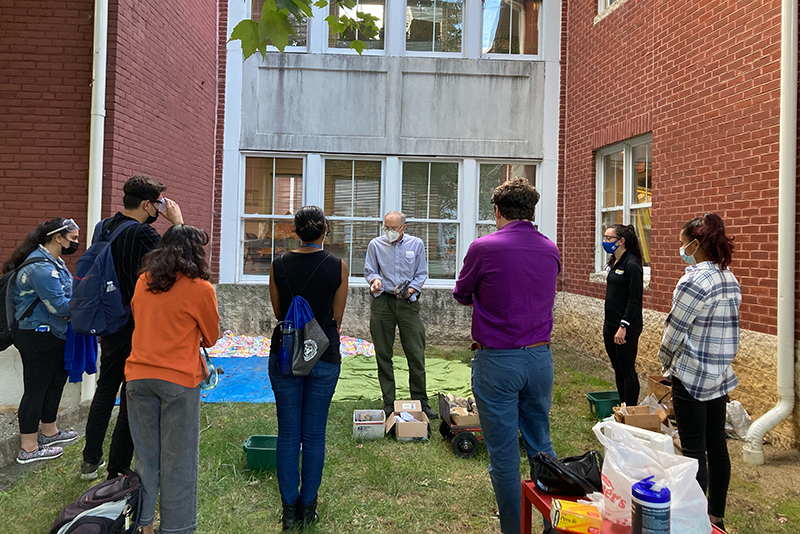 A UD professor stands outside next to a red brick building and demonstrates how to make stone tools as a group of college students observe.