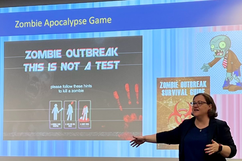 Professor Jennifer Trivedi lecturing about disaster preparedness in front of a projection screen with the words "zombie breakout this is not a test"