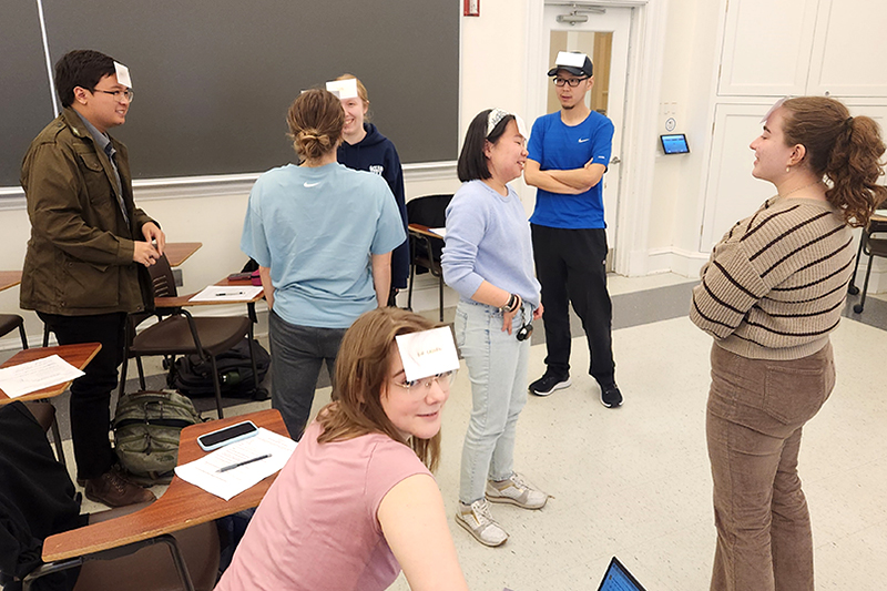 Several college students inside a classroom play a game with index cards taped to their foreheads.