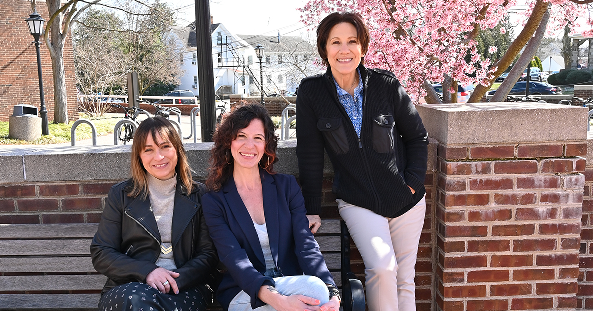 A group of three female University of Delaware professors on the UD campus in front of a red brick wall and a blooming pink cherry tree.