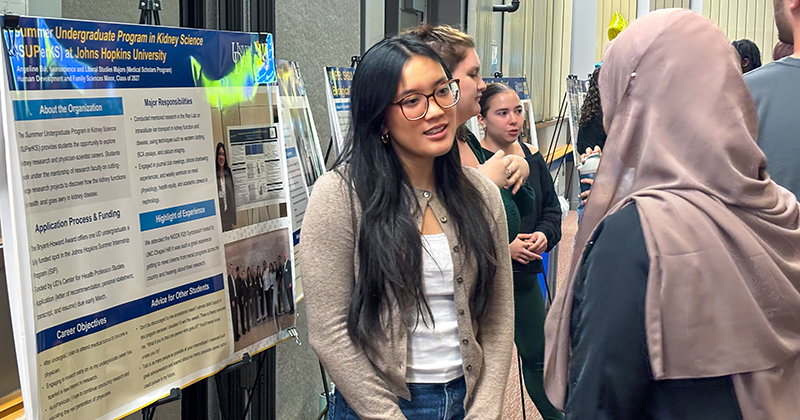 Poster presentation at Student Success Center internship symposium A college student at an event talks to another student while standing next to a poster on an easel.