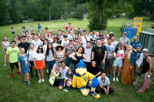 A group photo of about 60 international students and faculty with the YoUDee mascot standing on a lawn during a picnic.