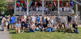 A group photo of about 60 international students standing on the porch and the lawn in front of a historic academic building decorated with colorful national flags.