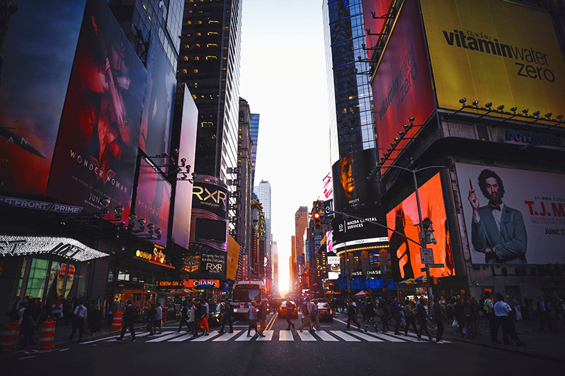 A crowd of people cross a busy street at sunset in New York City, surrounded by tall buildings.