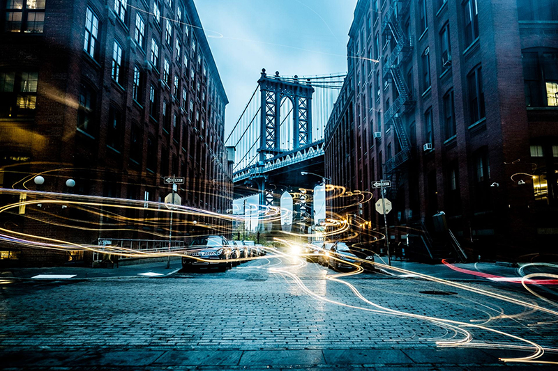 Cobblestone street intersection in New York City flanked by tall brick buildings with a view of a suspension bridge in the center.