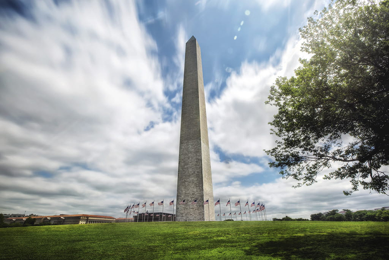 tall white obelisk on green lawn encircled by 50 American flags on flagpoles.