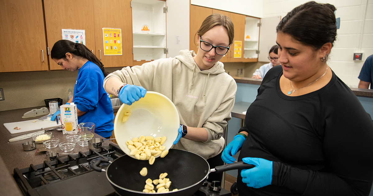 Students learn a healthier way to make gnocchi. Shown here, students are pouring gnocchi from a bowl into a frying pan on the stove in the Food and Nutrition Education Lab.