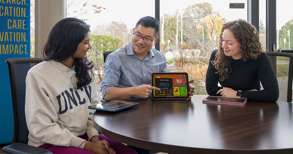 Daehyoung Lee, assistant professor of health behavior and nutrition sciences, sits at a round table and holds up an iPad, showing a 2,921 step-count on the PuzzleWalk app while a research assistant and doctoral student look at the app.