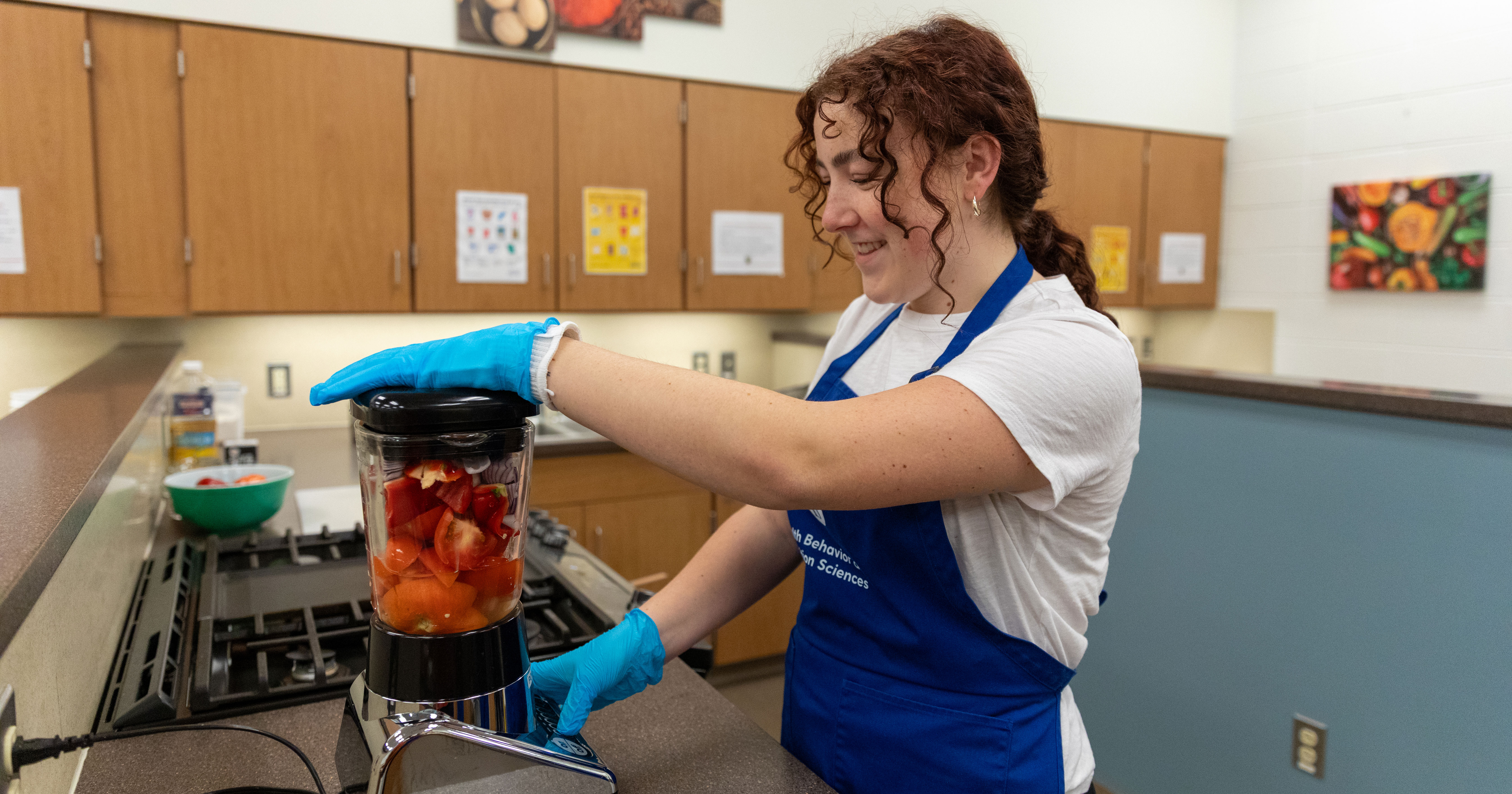 Celeste Phillips, a sophomore honors nutrition and dietetics major, wears a blue aprove and blue latex gloves as she places one hand on top of a blender, containing tomatos, and her other hand on the blend button, as a step in preparing jollof rice, a traditional Nigerian dish, for her Food Concepts course.