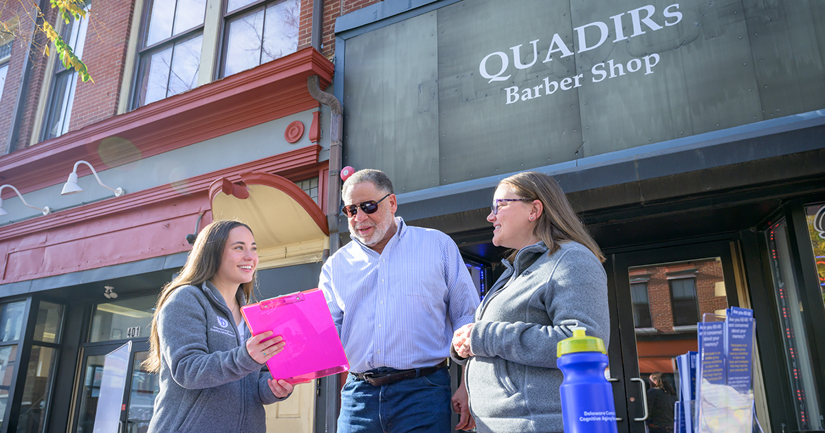 Olivia Dallas (left) and Faith Stagge (right), communication sciences and disorders doctoral students, set up shop outside Quadir’s Barber Shop in Wilmington as part of a novel, community-based strategy to recruit diverse study participants. Here, Olivia is holding a pink folder with paperwork while recruiting a study participant. Quadir's Barber Shop storefront on Market Street in Wilmington is shown in the background.