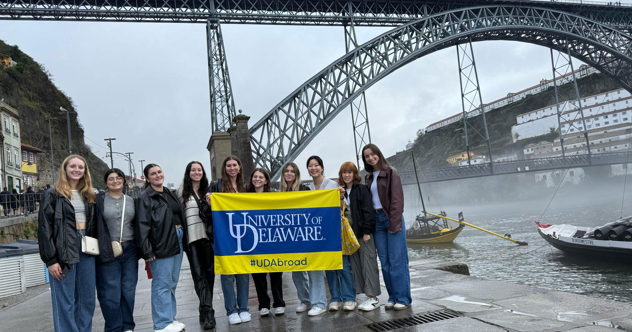 Students on a study abroad trip pose outside the most famous bridge in Portugal, the Dom Luis I Bridge, on a weekend excursion to Porto, holding a blue and yellow University of Delaware flag.