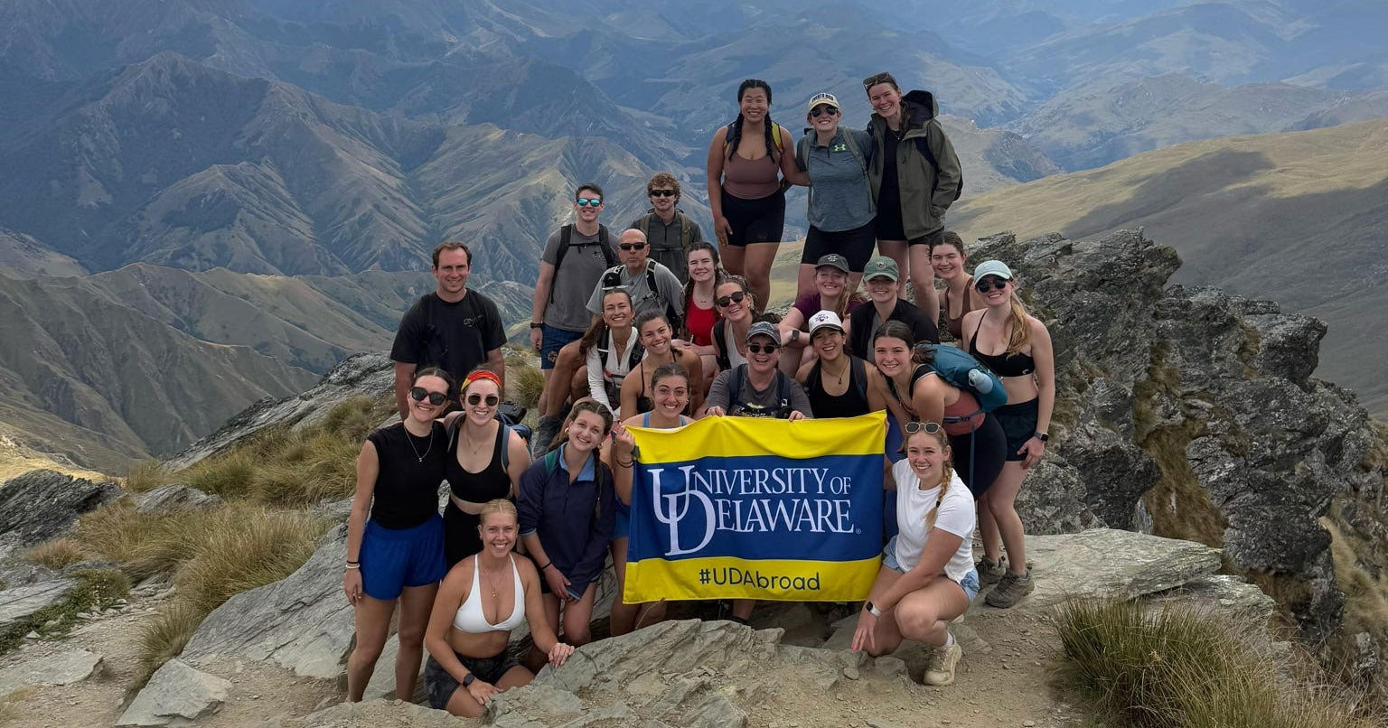 Students pose at the summit of Ben Lomond near Queenstown, New Zealand holding a UD blue and yellow flag. 