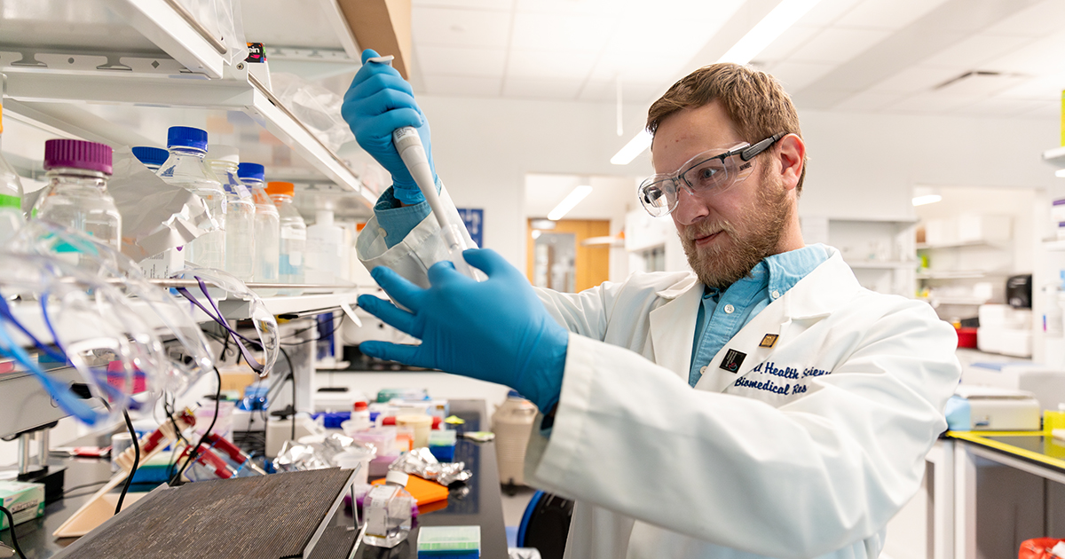 Ibra Fancher, assistant professor of kinesiology and applied physiology, works in a lab wearing a white coat, blue gloves and goggles. Here, he's shown holding a pipette in the air.