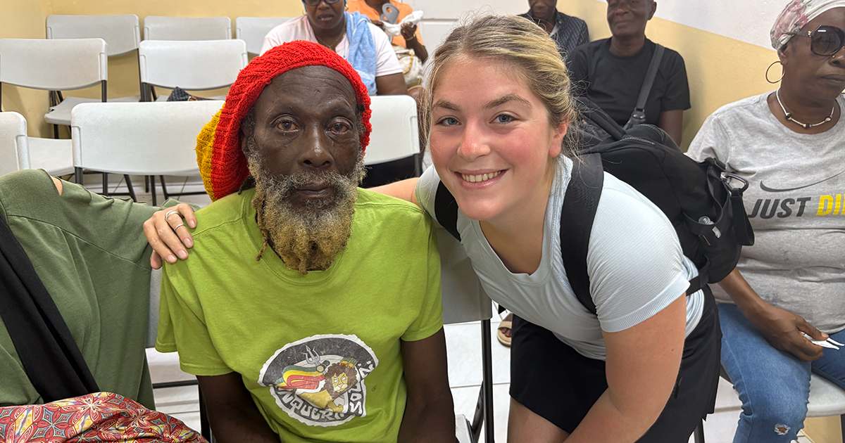 Senior medical diagnostics pre-physician assistant major Shayna Fink crouches down wearing a backpack on her shoulder and places her arm around a patient who recently had cataract surgery at a Vision Health International clinic during a volunteer medical  mission trip in Grenada.