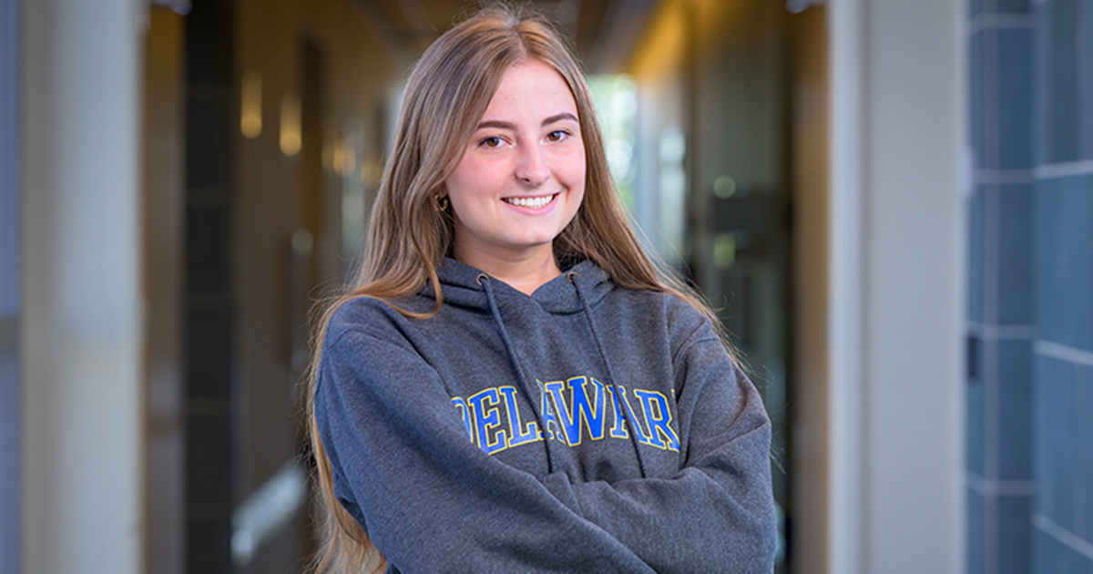 Gabriella Trincia wears a gray and blue UD sweatshirt and poses with her arms crossed in a building on campus with a hallway behind her.