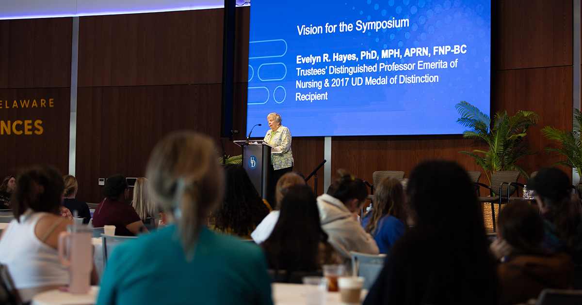 Evelyn R. Hayes, Trustees Distinguished Professor Emerita in UD’s School of Nursing, whose generous support makes the annual Hayes Symposium possible, delivers her vision for this year's symposium in a room full of people seated at round tables in the Audion on STAR Campus. Behind Evelyn is a large projection screen with a blue background that reads 