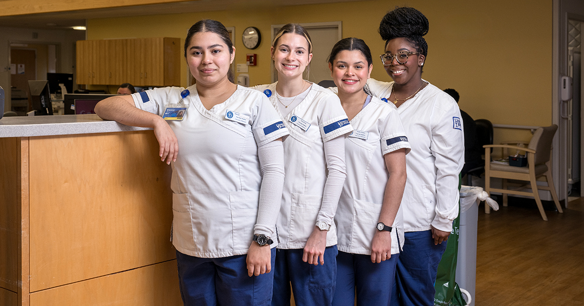 Four University of Delaware seniors have been named UD School of Nursing Bayhealth Scholars. Pictured here, they're wear their white and blue UD nursing scrubs and stand in a row in front of a wood nurses' station at Bayhealth Hospital, Kent Campus, where they'll complete their senior year clinicals and commit to working for two years.