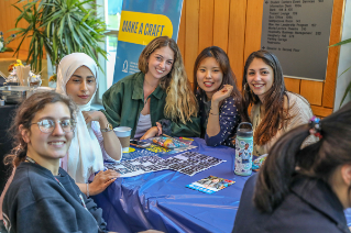 Five smiling college students sitting around an activities table during an event.