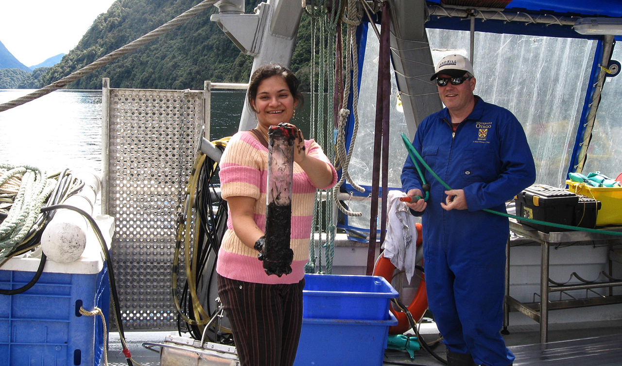 A Fulbright Faculty Scholar on a boat holding what looks to be a glass container that holds some sort of soil and water mixture.