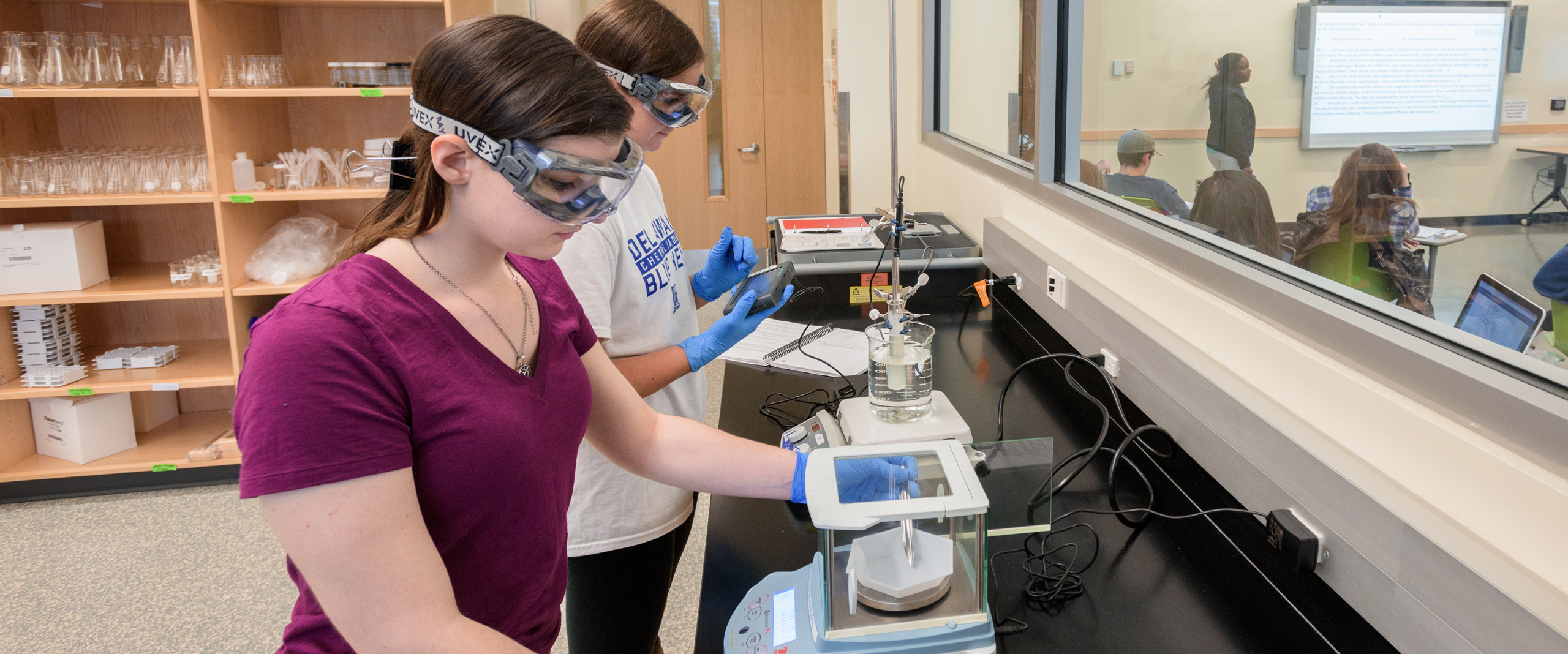 two students work with lab equipment in ISE while a neighboring classroom attends to a lecture