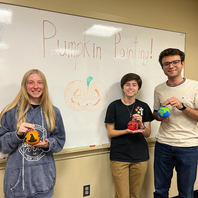 Students holding painted pumpkins in front of a whiteboard that read's "Pumpkin Painting"