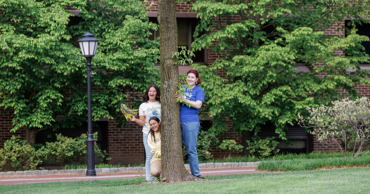 Three Student LIfe Orientation Leaders stand in a circle around a tree on campus wearing UD gear