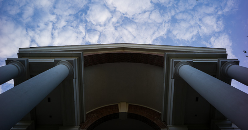 Camera looks up at the pillars of a large building with a blue sky and clouds.