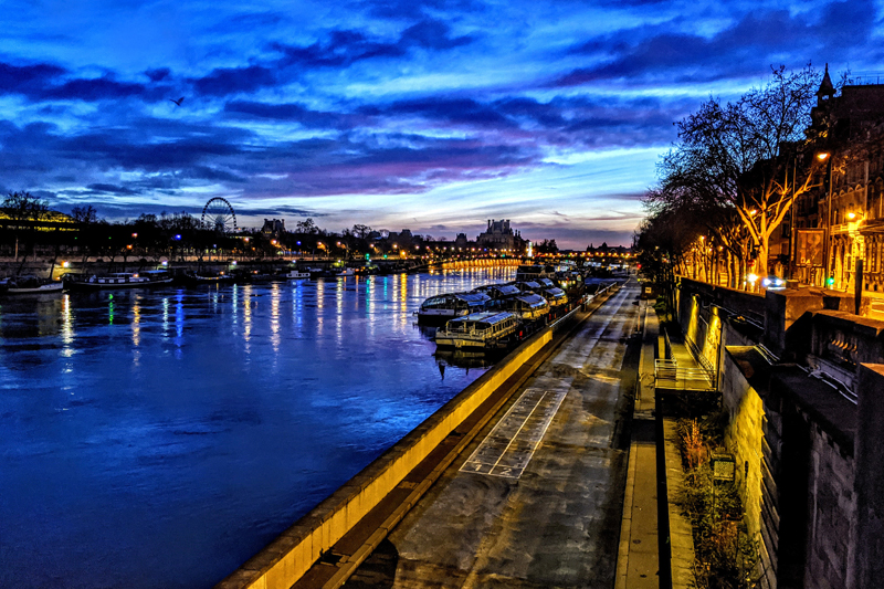 Across the Seine, and silhouetted against the brightening early morning sky, is the Louvre museum in the distance and the Ferris Wheel of the La Magie de Noël Christmas festival. 