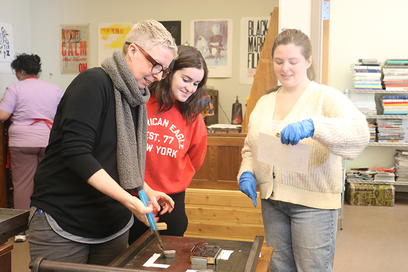 Associate Professor of English Laura Helton shows Gia Joella and Cassidy Driscoll how to place tiles on the hand press. If the tiles move when the roller is pulled across, the printed image and letters will smear. Associate Professor of English Laura Helton shows Gia Joella and Cassidy Driscoll how to place tiles on the hand press. If the tiles move when the roller is pulled across, the printed image and letters will smear.